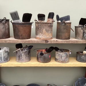 Op de afbeelding: Een verzameling verweerde metalen emmers en bakjes op twee houten planken. Elke houder bevat een of meer verfkwasten met donkere haren. De metalen oppervlakken vertonen sporen van roest, slijtage en verfspatten in diverse tinten.