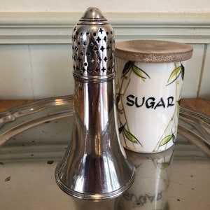 May include: A silver sugar shaker with a perforated top and a bell-shaped base. The shaker is sitting on a silver tray next to a white ceramic sugar jar with a wooden lid and the word "SUGAR" written on it.