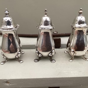 May include: Three antique silver salt and pepper shakers. Each shaker has a bulbous body, a perforated lid, and rests on three small feet. The shakers are highly reflective, with a polished finish, and are arranged on a light-colored surface.