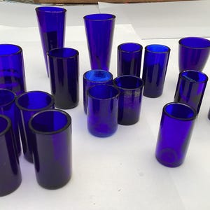 Könnte beinhalten: Eine Auswahl an Trinkgläsern aus kobaltblauem Glas in verschiedenen Formen und Größen. Die Kollektion umfasst hohe, zylindrische und konische Designs, alle mit glänzendem Finish. Die Gläser sind auf einer weißen Oberfläche angeordnet und zeigen ihre lebendige Farbe und reflektierenden Eigenschaften.