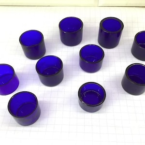 Puede incluir: Una colección de diez portavelas votivas de vidrio azul cobalto. Cada soporte cilíndrico tiene un borde oscuro y un interior azul intenso, creando un contraste llamativo. Los soportes están dispuestos sobre una superficie de cuadrícula blanca.