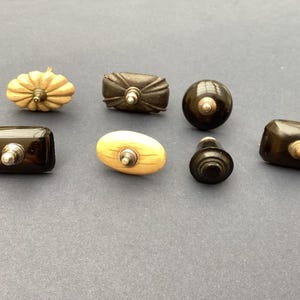 May include: A selection of seven antique cabinet knobs in different shapes and materials. The knobs include a pumpkin-shaped, a rectangular, and a round knob, all with metal fixings. The colours vary from light beige to dark brown.