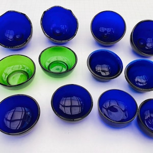 Könnte beinhalten: Eine Auswahl kleiner, runder Glasschalen in Blau- und Grüntönen. Die Schalen haben einen dunklen Rand und eine glatte, reflektierende Oberfläche. Die Schalen sind in einer Gruppe angeordnet und zeigen ihre unterschiedlichen Farben und Formen.