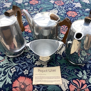 May include: A collection of Picquot Ware tea and coffee service items. The set includes a teapot, coffee pot, creamer, and a lidded coffee server. Each piece is made of polished aluminium with wooden handles and accents. A cleaning instructions card is also present.