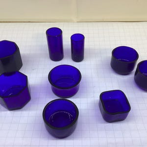 May include: An assortment of cobalt blue glass containers in various shapes. The collection includes cylindrical, hexagonal, and rounded forms, some with a black rim. The containers are arranged on a white surface.