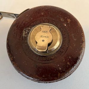 Puede incluir: Una cinta métrica vintage cubierta de cuero con un mecanismo de bobinado de latón. El mecanismo tiene el texto "66 FT" y "N9418" grabado.