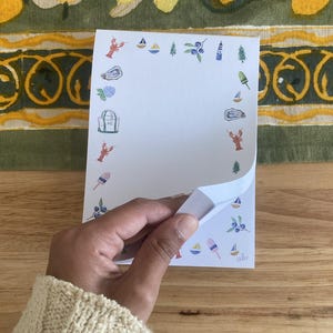 May include: A white notepad with a blue and red border. The border is decorated with small illustrations of sailboats, lobsters, blueberries, lighthouses, and ice cream cones.