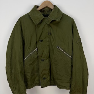 May include: An olive green jacket featuring a wide collar, a front button closure, and two diagonal zippered pockets on the chest. The jacket has long sleeves with adjustable cuffs and a utility pocket on the left sleeve, hanging on a wooden hanger.