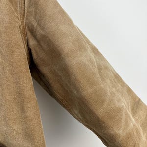 Puede incluir: Primer plano de una manga de chaqueta de trabajo color beige. La chaqueta est&aacute; hecha de una tela duradera y texturizada con costuras visibles a lo largo de las costuras. La manga est&aacute; ligeramente desgastada, mostrando signos de uso. La chaqueta tiene un bolsillo en la parte inferior izquierda.