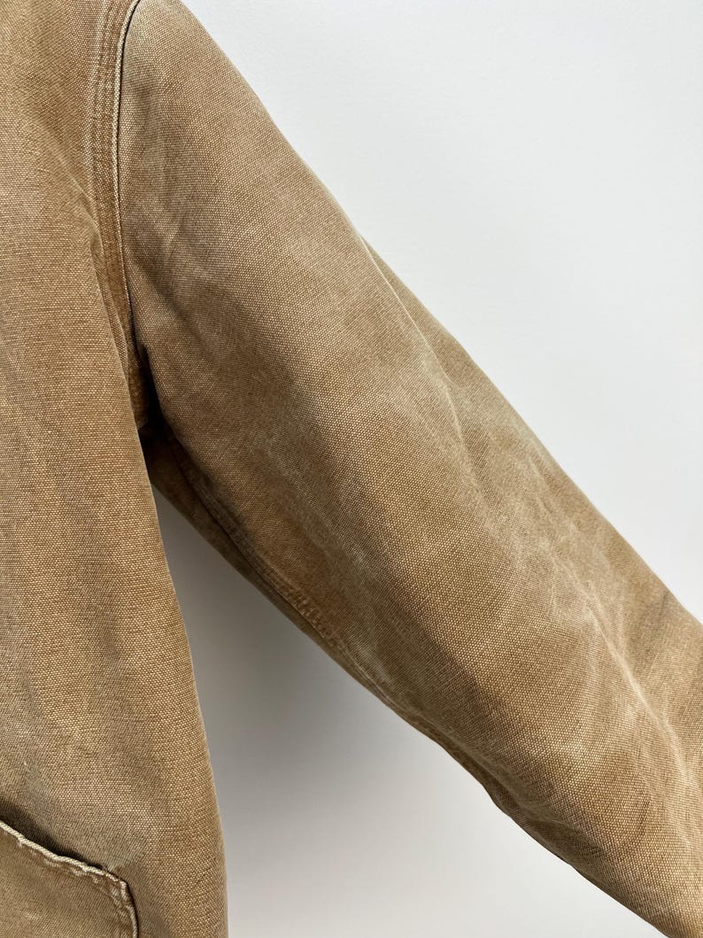 Puede incluir: Primer plano de una manga de chaqueta de trabajo color beige. La chaqueta est&aacute; hecha de una tela duradera y texturizada con costuras visibles a lo largo de las costuras. La manga est&aacute; ligeramente desgastada, mostrando signos de uso. La chaqueta tiene un bolsillo en la parte inferior izquierda.