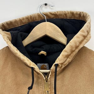 Puede incluir: Primer plano de una chaqueta con capucha de color canela con forro acolchado negro. La chaqueta tiene una cremallera completa, cordones negros y un peque&ntilde;o parche con el logotipo. La capucha est&aacute; forrada con tela negra y cuelga de una percha de madera.