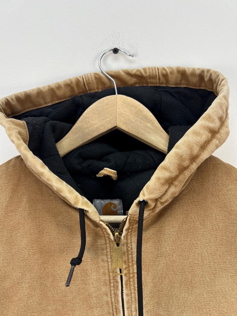 Puede incluir: Primer plano de una chaqueta con capucha de color canela con forro acolchado negro. La chaqueta tiene una cremallera completa, cordones negros y un peque&ntilde;o parche con el logotipo. La capucha est&aacute; forrada con tela negra y cuelga de una percha de madera.