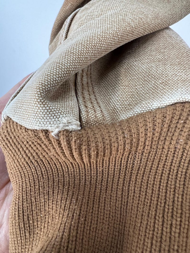 Puede incluir: Primer plano de una tela beige con textura tejida, superpuesta a un material de punto marr&oacute;n acanalado. Las telas est&aacute;n dobladas, revelando las diferentes texturas y colores. La imagen resalta el contraste entre el material tejido liso y el punto texturizado.