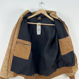 Puede incluir: Chaqueta Carhartt marr&oacute;n con forro acolchado negro, expuesta abierta en una percha de madera. La chaqueta tiene m&uacute;ltiples bolsillos, capucha y dobladillo y pu&ntilde;os acanalados. El exterior es de color marr&oacute;n claro.