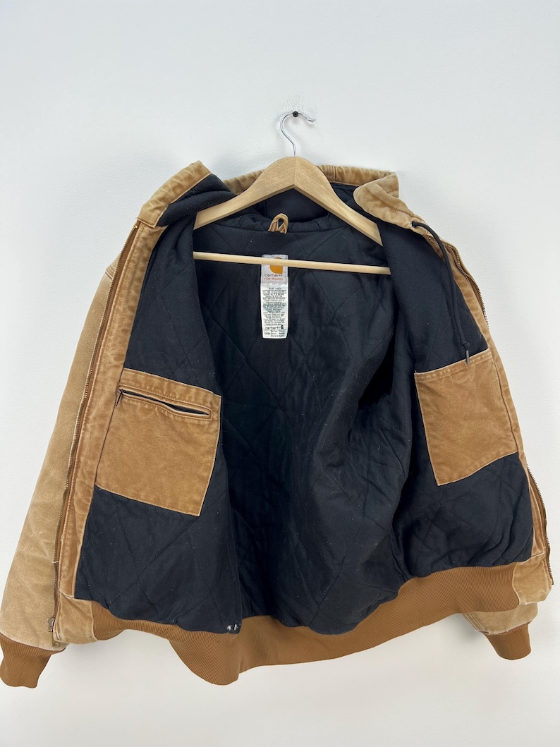 Puede incluir: Chaqueta Carhartt marr&oacute;n con forro acolchado negro, expuesta abierta en una percha de madera. La chaqueta tiene m&uacute;ltiples bolsillos, capucha y dobladillo y pu&ntilde;os acanalados. El exterior es de color marr&oacute;n claro.