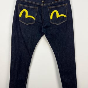 Op de afbeelding: Donkerblauwe denim jeans met gele geschilderde details op de achterzakken. De jeans hebben een klassiek vijf-zakken ontwerp, een bruine leren patch en een knoop- en ritssluiting.