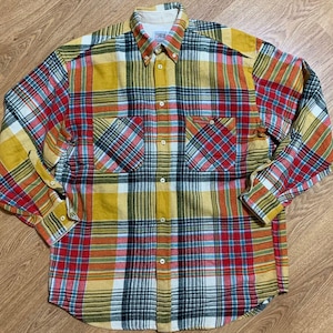 May include: A yellow, red, blue and black plaid flannel shirt with a button-up front and long sleeves. The shirt has two chest pockets and a brand label that reads "Timberland" on the left side of the collar.