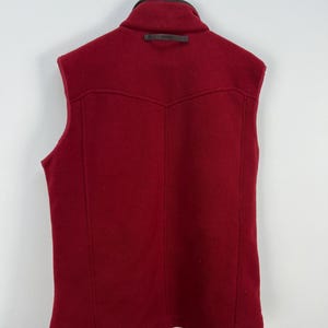 May include: A sleeveless, red vest with a high collar and a leather accent. The back of the vest features a yoke design and vertical seams. The vest is hanging on a metal hanger against a white background.