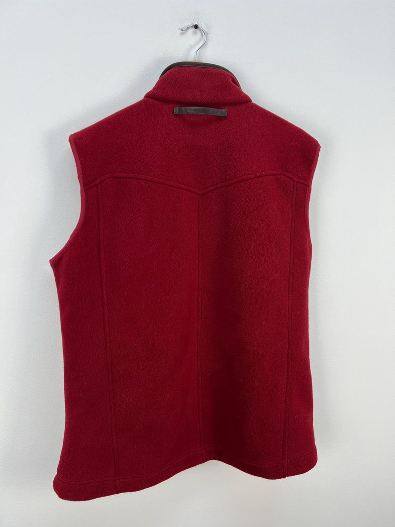 May include: A sleeveless, red vest with a high collar and a leather accent. The back of the vest features a yoke design and vertical seams. The vest is hanging on a metal hanger against a white background.