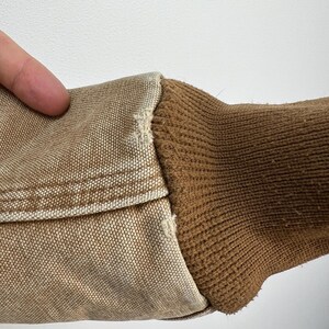 Puede incluir: Primer plano de una manga de chaqueta de trabajo color beige. La manga presenta un pu&ntilde;o de punto acanalado marr&oacute;n y una tela tipo lona beige. Las costuras son visibles, mostrando la construcci&oacute;n de la prenda. El fondo es una superficie blanca lisa.