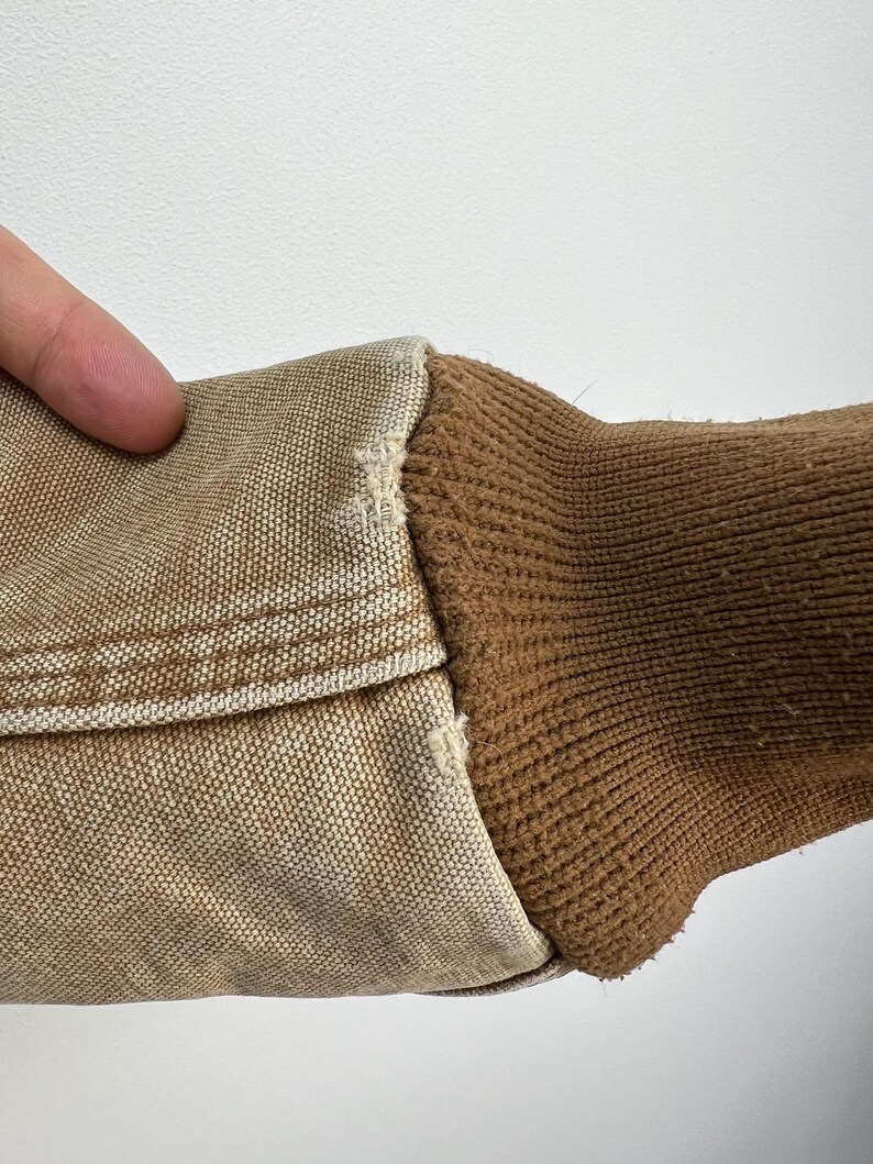 Puede incluir: Primer plano de una manga de chaqueta de trabajo color beige. La manga presenta un pu&ntilde;o de punto acanalado marr&oacute;n y una tela tipo lona beige. Las costuras son visibles, mostrando la construcci&oacute;n de la prenda. El fondo es una superficie blanca lisa.