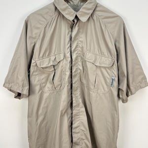 May include: A tan short-sleeved shirt with two chest pockets and a collared neckline. The shirt is made of a lightweight material and is hanging on a wooden hanger. The shirt has a button-down front.