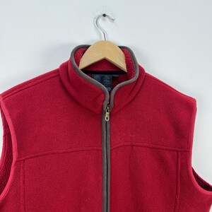 May include: A red fleece vest with a full-length zipper and a brown trim around the collar and armholes. The vest is hanging on a wooden hanger against a white background. The vest has a soft, textured appearance.