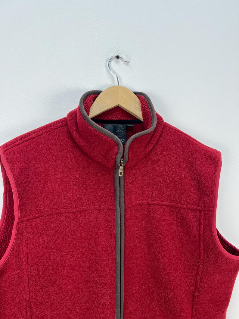 May include: A red fleece vest with a full-length zipper and a brown trim around the collar and armholes. The vest is hanging on a wooden hanger against a white background. The vest has a soft, textured appearance.