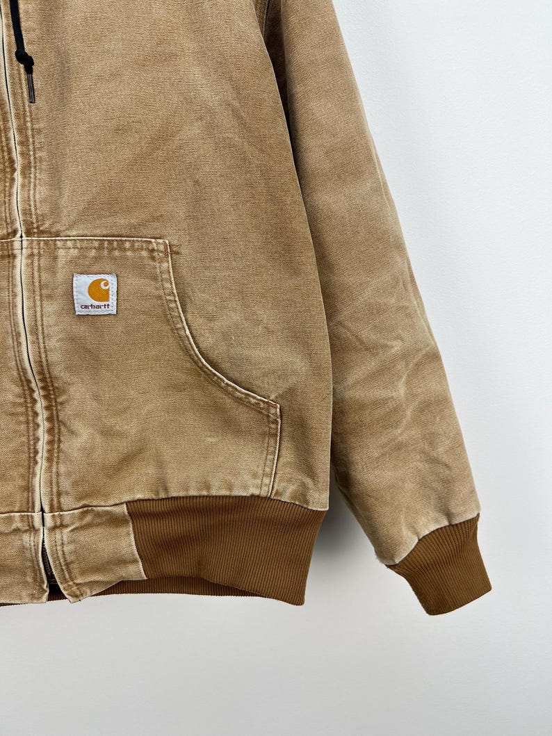 Puede incluir: Chaqueta con capucha Carhartt de color marr&oacute;n claro con cremallera frontal, bolsillo tipo canguro y pu&ntilde;os y dobladillo acanalados. El logotipo de Carhartt est&aacute; en un peque&ntilde;o parche cuadrado en el bolsillo delantero. Tejido resistente.