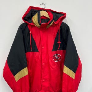 Könnte beinhalten: Rote, schwarze und goldene Kapuzenjacke mit einem San Francisco 49ers Logo auf der Vorderseite. Die Jacke hat einen Reißverschluss und eine Kapuze mit Kordelzug.