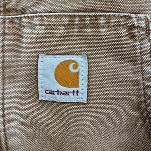 Puede incluir: Primer plano de una prenda de trabajo marr&oacute;n Carhartt, que muestra el parche con el logotipo de la marca. El parche es rectangular con fondo blanco y el logotipo de Carhartt en naranja y blanco. La tela tiene un aspecto texturizado.