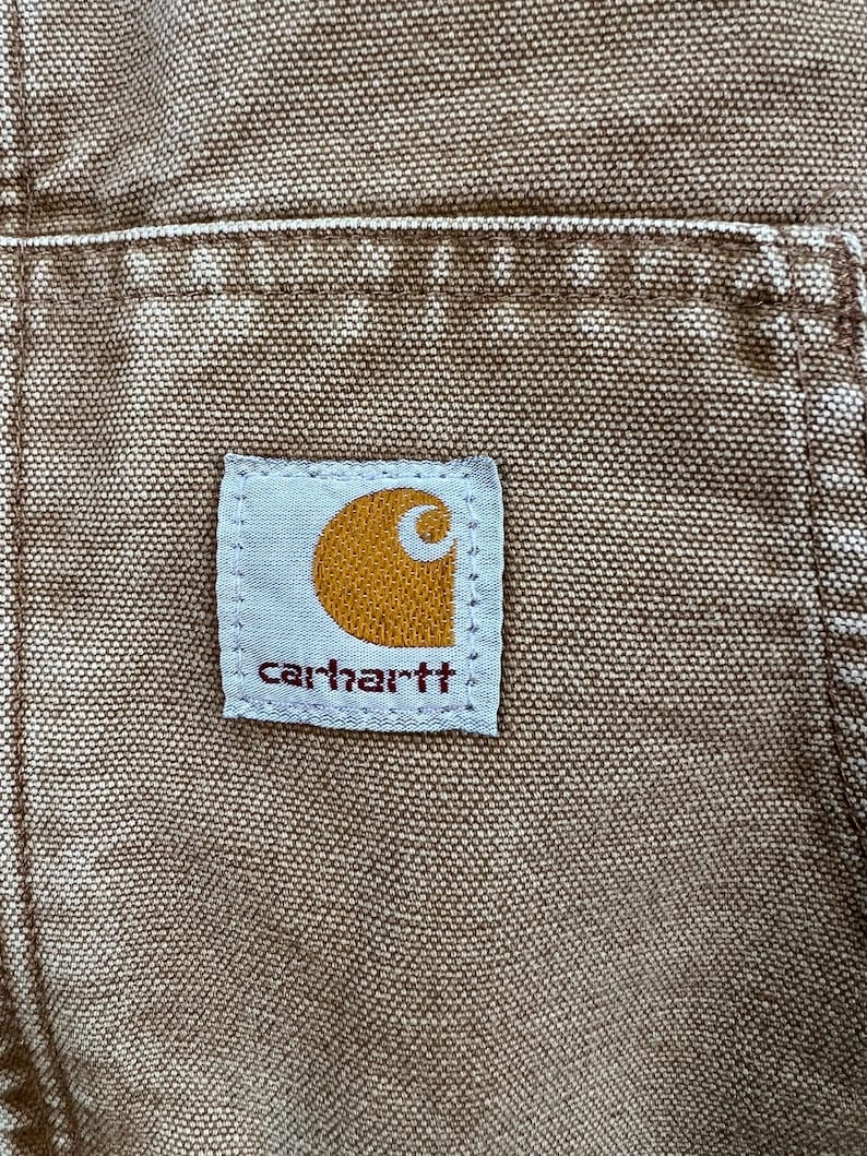 Puede incluir: Primer plano de una prenda de trabajo marr&oacute;n Carhartt, que muestra el parche con el logotipo de la marca. El parche es rectangular con fondo blanco y el logotipo de Carhartt en naranja y blanco. La tela tiene un aspecto texturizado.
