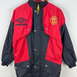 Könnte beinhalten: Rote und schwarze Vintage Umbro Manchester United Jacke. Die Jacke zeigt das Umbro-Logo und das Manchester United-Wappen. Sie hat einen gelben Kragen und einen Reißverschluss. Die Jacke hat eine Knopfleiste und zwei Taschen.