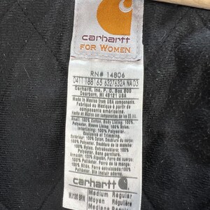Puede incluir: Primer plano de una etiqueta Carhartt, con el logotipo de la marca y el texto "FOR WOMEN". La etiqueta proporciona detalles sobre los materiales y las instrucciones de cuidado de la prenda. El fondo es una tela oscura y acolchada.