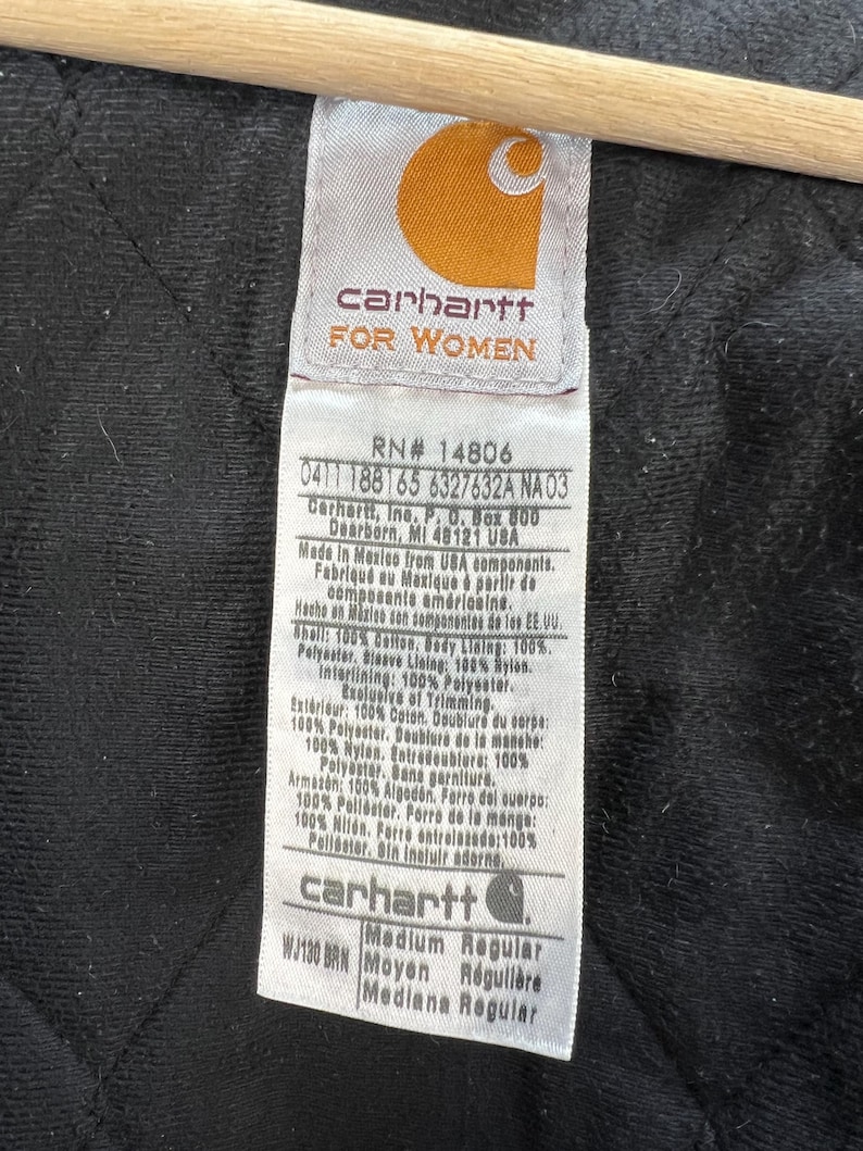 Puede incluir: Primer plano de una etiqueta Carhartt, con el logotipo de la marca y el texto "FOR WOMEN". La etiqueta proporciona detalles sobre los materiales y las instrucciones de cuidado de la prenda. El fondo es una tela oscura y acolchada.