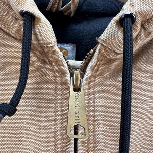 Puede incluir: Primer plano de una chaqueta con capucha Carhartt de color canela. La chaqueta presenta una cremallera de lat&oacute;n con el logotipo de Carhartt, cordones negros y un forro interior negro. La capucha es visible y la tela tiene un aspecto texturizado.