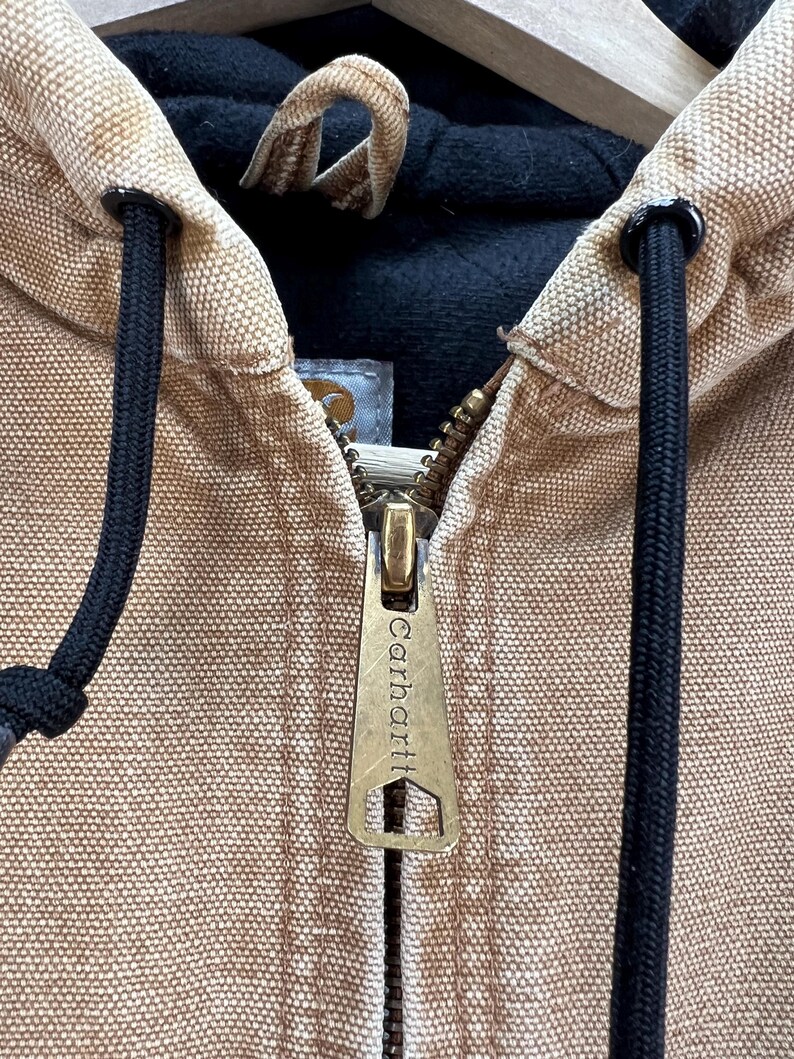 Puede incluir: Primer plano de una chaqueta con capucha Carhartt de color canela. La chaqueta presenta una cremallera de lat&oacute;n con el logotipo de Carhartt, cordones negros y un forro interior negro. La capucha es visible y la tela tiene un aspecto texturizado.