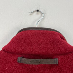 May include: Close-up of a red fleece jacket collar with a brown trim and a brown label that reads "MUSTO". The jacket is hanging on a silver hanger against a white background. The fleece has a textured appearance.