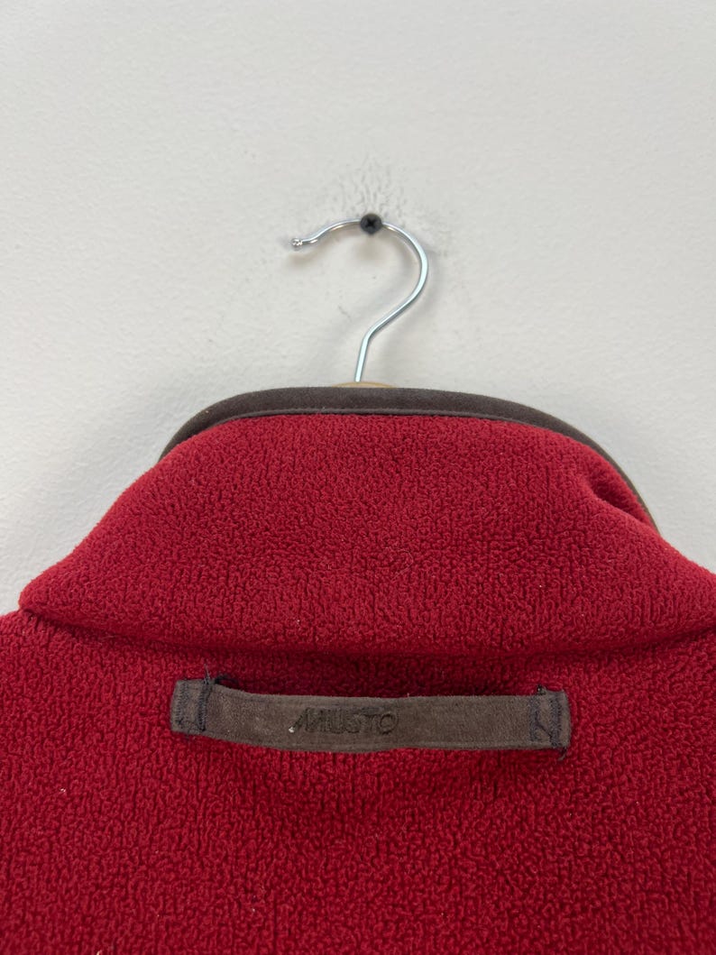May include: Close-up of a red fleece jacket collar with a brown trim and a brown label that reads "MUSTO". The jacket is hanging on a silver hanger against a white background. The fleece has a textured appearance.