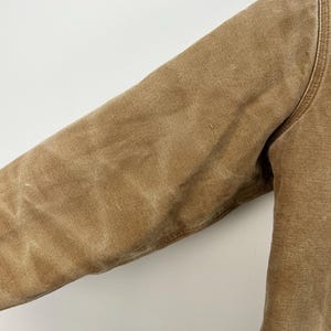 Puede incluir: Primer plano de una manga de chaqueta de trabajo color canela. La chaqueta est&aacute; hecha de una tela duradera y texturizada. La manga es de color canela liso con costuras visibles a lo largo de las costuras. La chaqueta parece estar bien usada.