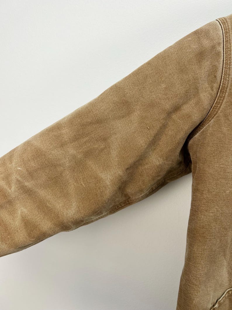 Puede incluir: Primer plano de una manga de chaqueta de trabajo color canela. La chaqueta est&aacute; hecha de una tela duradera y texturizada. La manga es de color canela liso con costuras visibles a lo largo de las costuras. La chaqueta parece estar bien usada.