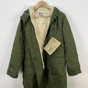 Puede incluir: Parka verde oliva con forro de piel sintética color crema. La chaqueta tiene capucha, cierres de botón y múltiples bolsillos. Una etiqueta está adherida al frente. La parka está colgada de una percha de madera.