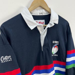 Pode incluir: Uma camisa de rugby preta com gola branca e listras multicoloridas vermelhas, azuis e verdes. A camisa tem o logotipo "Cotton Traders" na manga e o logotipo "RBS 6 Nations" no peito.