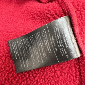 May include: Close-up of a textured red fabric with a black label. The label contains text about the fabric's composition, including polyester and cationic dyeable polyester. The brand name MUSTO is visible.