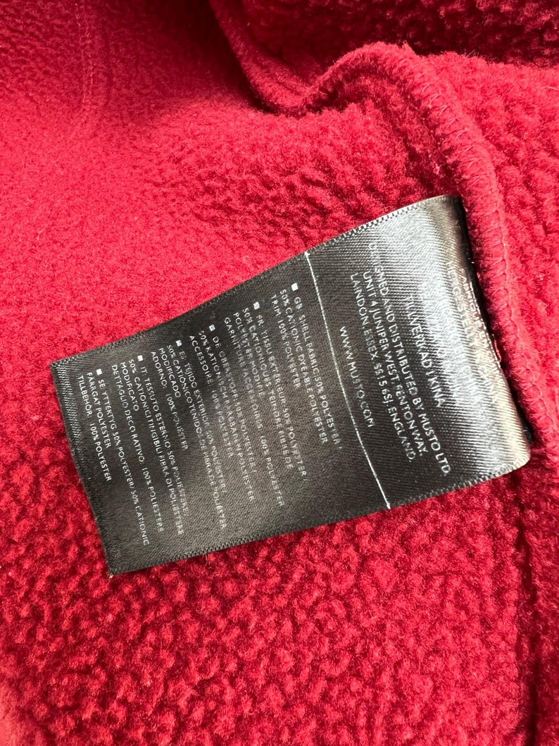 May include: Close-up of a textured red fabric with a black label. The label contains text about the fabric's composition, including polyester and cationic dyeable polyester. The brand name MUSTO is visible.