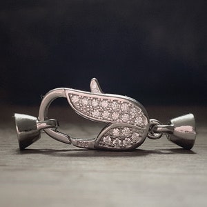 Könnte beinhalten: Silberfarbener Verschluss mit einem blattförmigen Design, das mit klaren Strasssteinen besetzt ist. Der Verschluss hat einen federbelasteten Mechanismus und eine kleine, runde Endkappe.