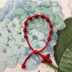 May include: A red knotted rosary bracelet with a cross pendant. The bracelet features multiple knots and a cross-shaped pendant. The background includes a floral design with blue and green hues.