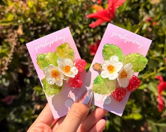 May include: Two hair clips featuring white flowers with orange centers, green leaves, and small red strawberries. Each clip is attached to a light pink display card with the text gemsbysidney printed in white script at the top.
