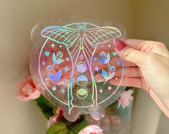 May include: A holographic sticker featuring a moth with a moon and floral design. The sticker is clear and has a rainbow iridescent finish.