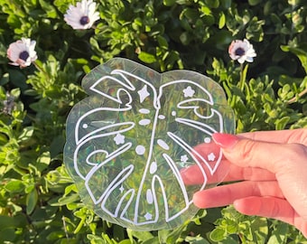 May include: A clear iridescent sticker with a white outline of a monstera leaf with stars inside. The sticker is being held in a hand.